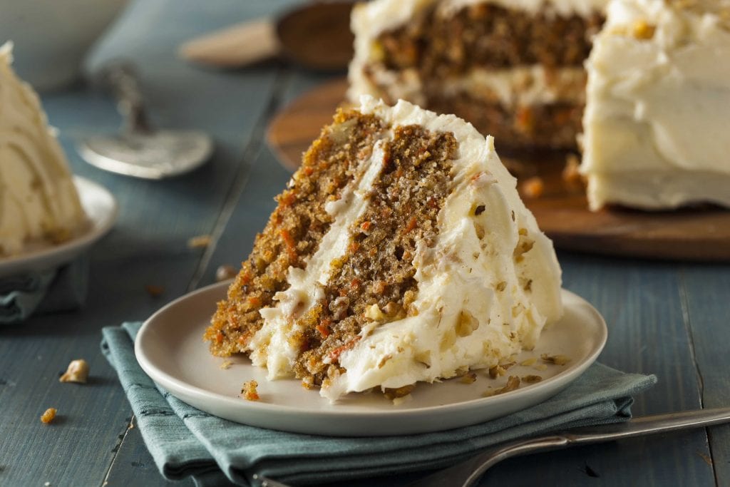 a slice of two-layered carrot cake covered in icing, with the full cake in the background