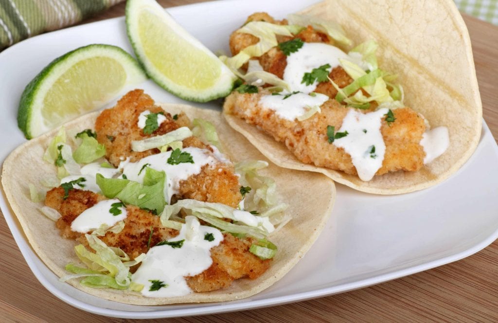2 fried fish tacos with 2 lime wedges on a white plate
