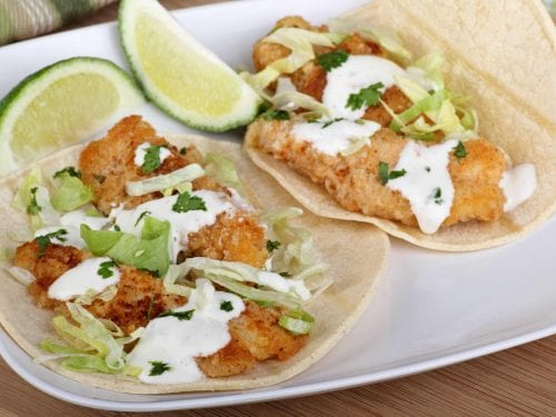 2 fried fish tacos with 2 lime wedges on a white plate