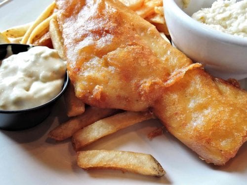 closeup on fish and chips on a white plate with 2 small sauce bowls of tartar sauce