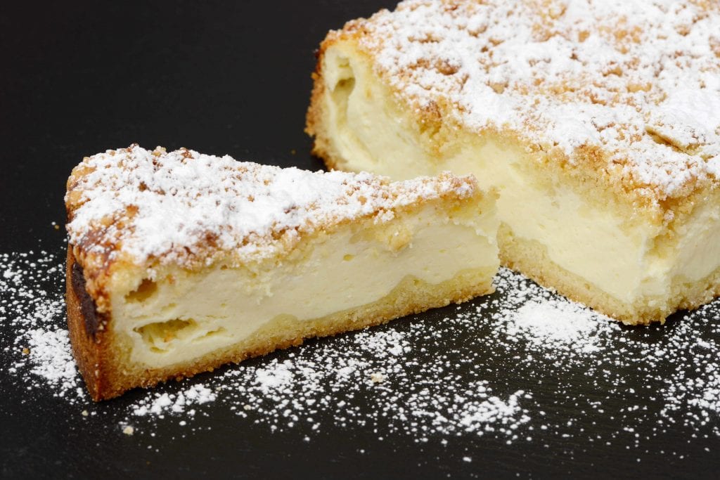 part of a cheesecake and one of the slices against a black background covered in powdered sugar