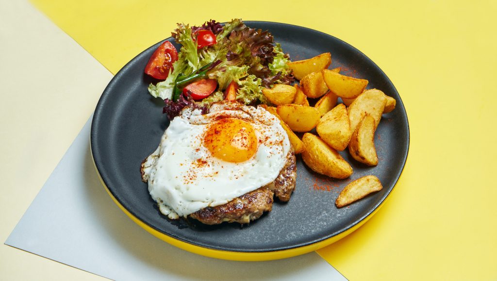 Beef steak with fried eggs with a side dish of salad and fried potatoes on a black plate on a colored background. Appetizing food for lunch