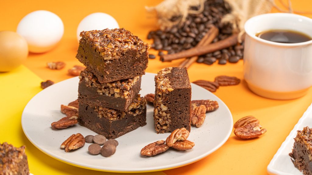 Pecan Pie Brownies Recipe, Pecan pie brownies cut into squares and served on top of a white plate decorated with pecans and chocolate chips