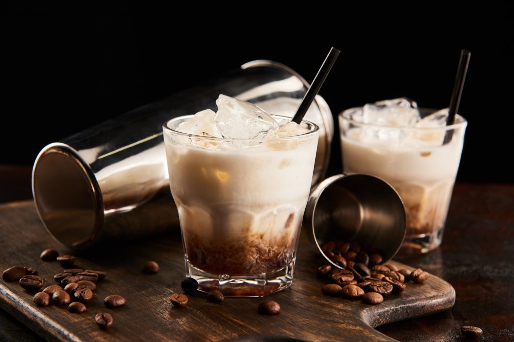 white russian cocktail in glasses with straws on wooden board with coffee grains and shaker isolated on black