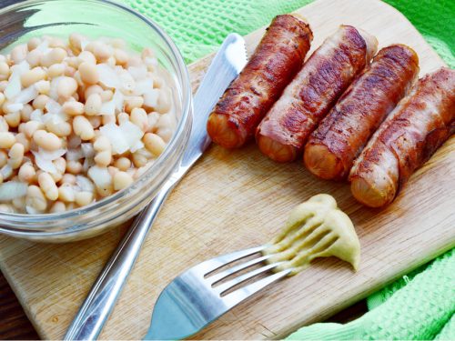Four pieces of grilled bacon-wrapped stuffed hot dogs, with a bowl of beans on the side.