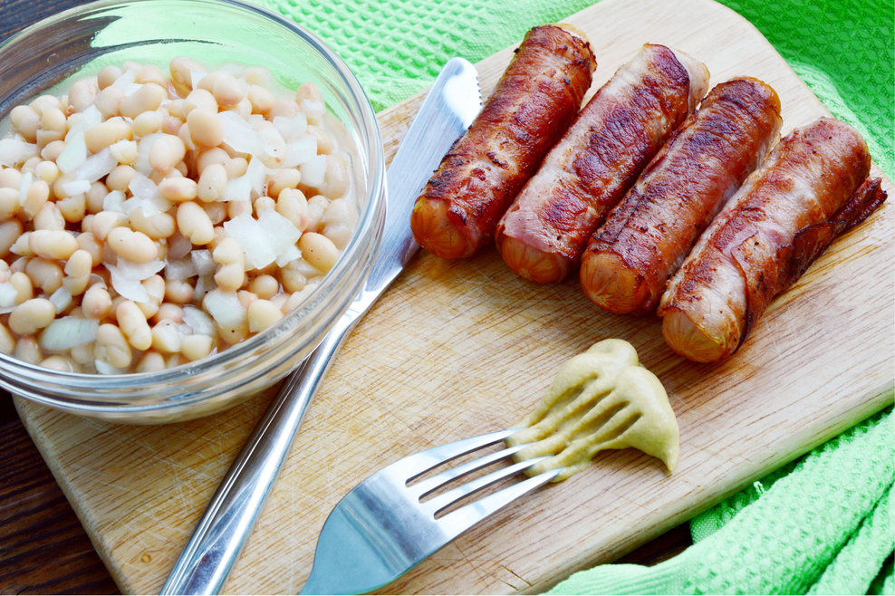 Four pieces of grilled bacon-wrapped stuffed hot dogs, with a bowl of beans on the side.