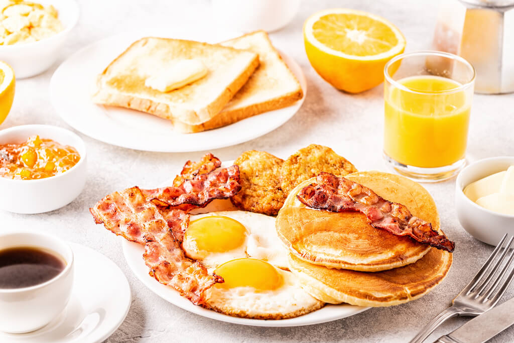 Full American breakfast, pancakes, bacon, eggs, toasts, orange juice, and black coffee on table