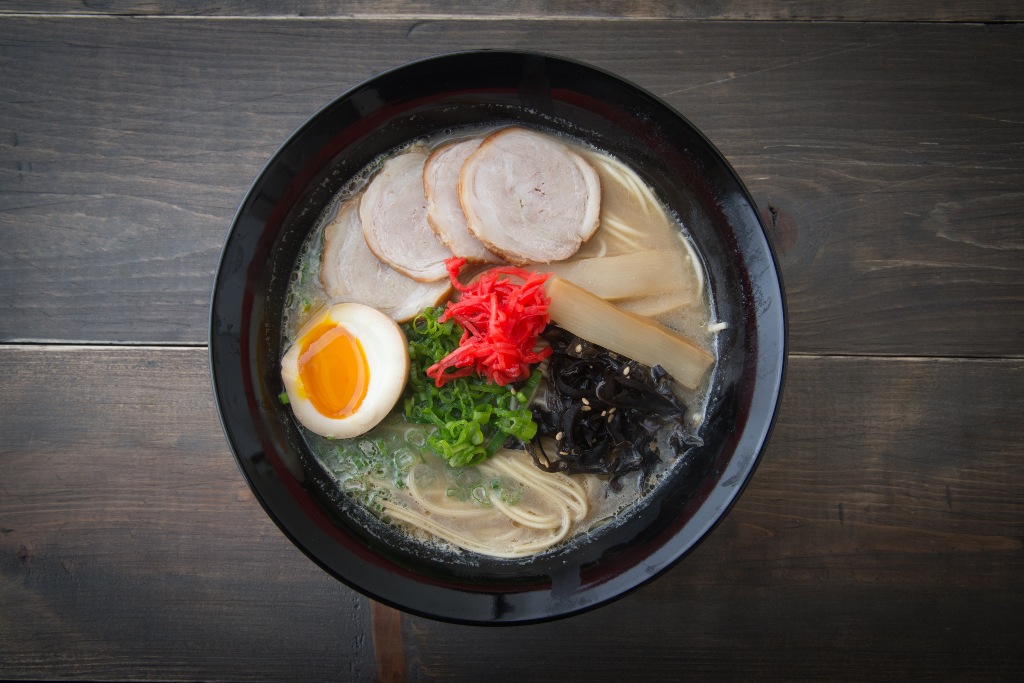 Bowl of Hakata-style ramen, with noodles in milky tonkotsu broth, topped with chashu, soft-boiled egg, fermented bamboo shoots, wood ear mushroom, pickled radish, chopped green onions, and sesame seeds