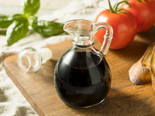 A bottle of balsamic vinegar on a wooden chopping board beside tomatoes, basil, and bread.