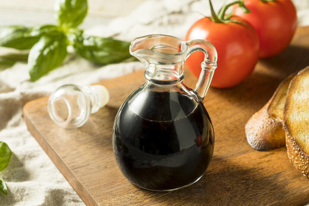 A bottle of balsamic vinegar on a wooden chopping board beside tomatoes, basil, and bread.